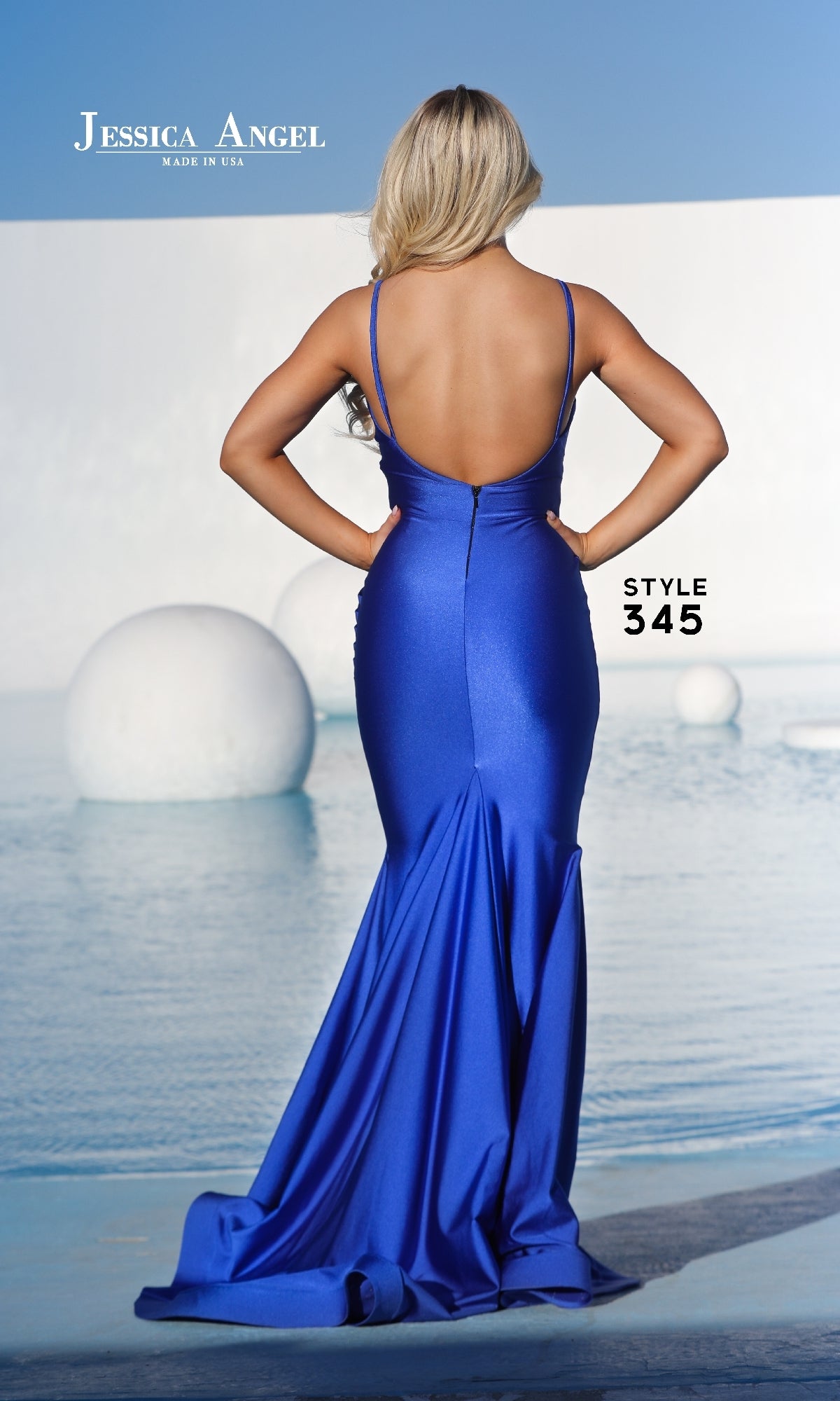 A woman with long blonde hair models the Jessica Angel 345 Long Glitter Mermaid Prom Dress—a backless, floor-length royal blue gown—posing by water with bold white geometric shapes behind her.